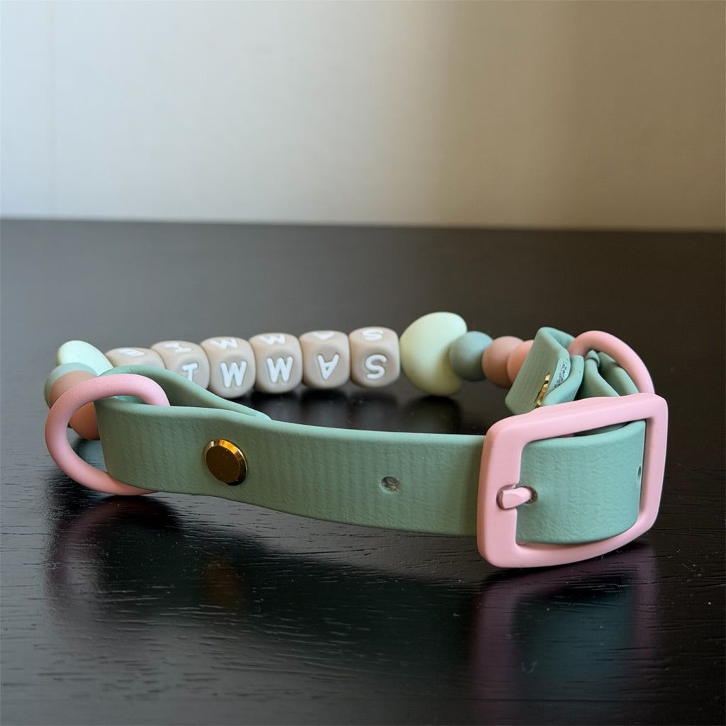 A dog collar made with biothane and silicone. The collar features a sage green biothane strap with pink rings and buckle, decorated with pastel pink, sage green and beige beads, and a bottom row of beige letter cubes spelling 'sammie'.