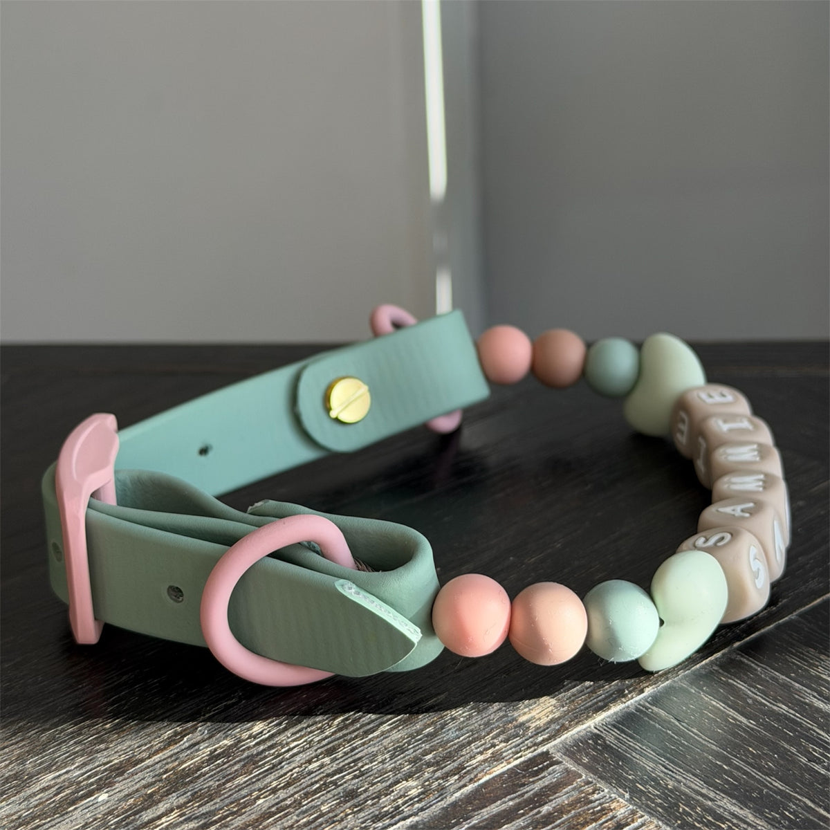 A dog collar made with biothane and silicone. The collar features a sage green biothane strap with pink rings and buckle, decorated with pastel pink, sage green and beige beads, and a bottom row of beige letter cubes spelling 'sammie'.