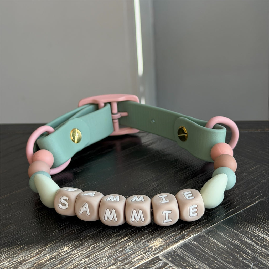A dog collar made with biothane and silicone. The collar features a sage green biothane strap with pink rings and buckle, decorated with pastel pink, sage green and beige beads, and a bottom row of beige letter cubes spelling 'sammie'.