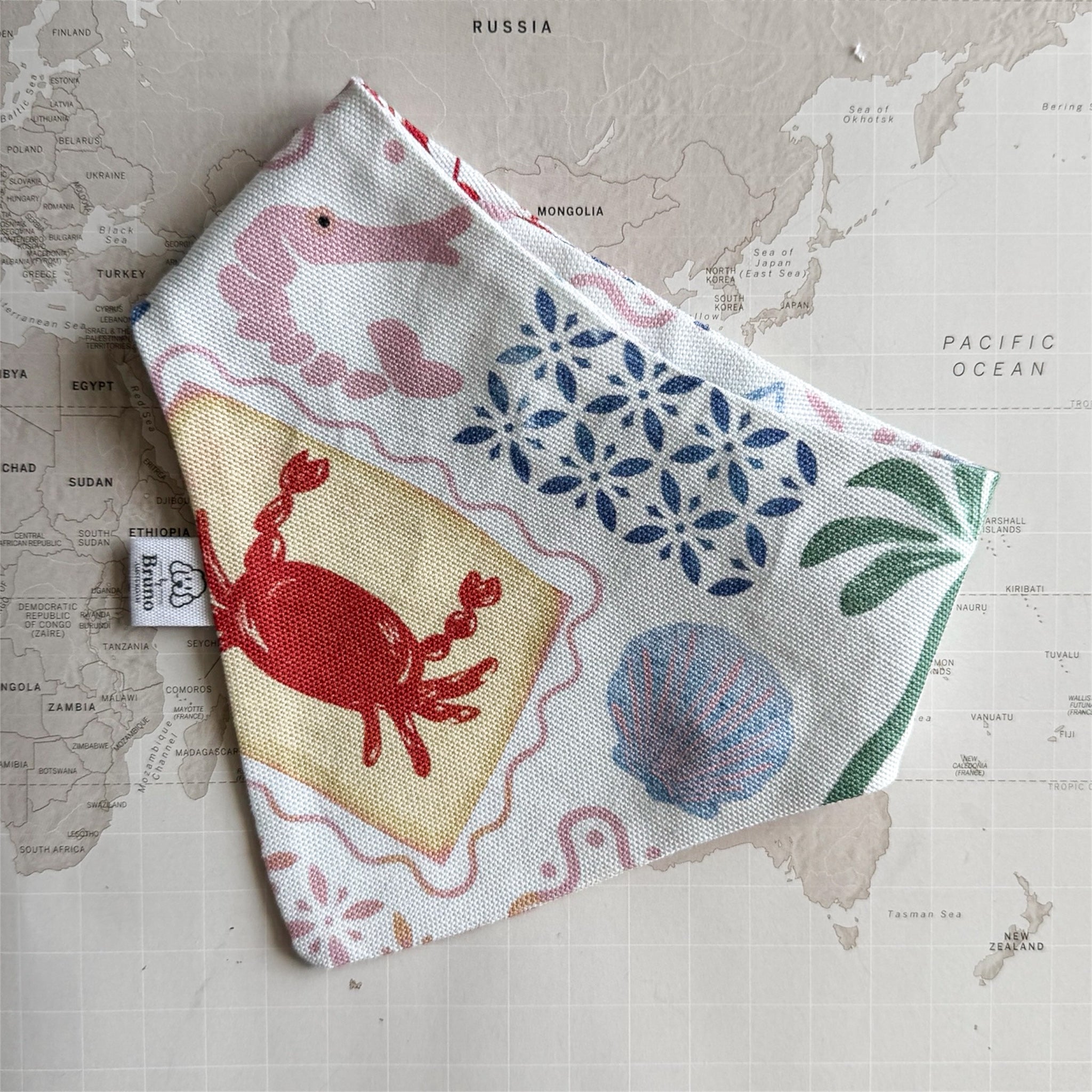 White handmade dog bandana with pastel summer symbols like palm trees, crab and sea shell on a world map background.