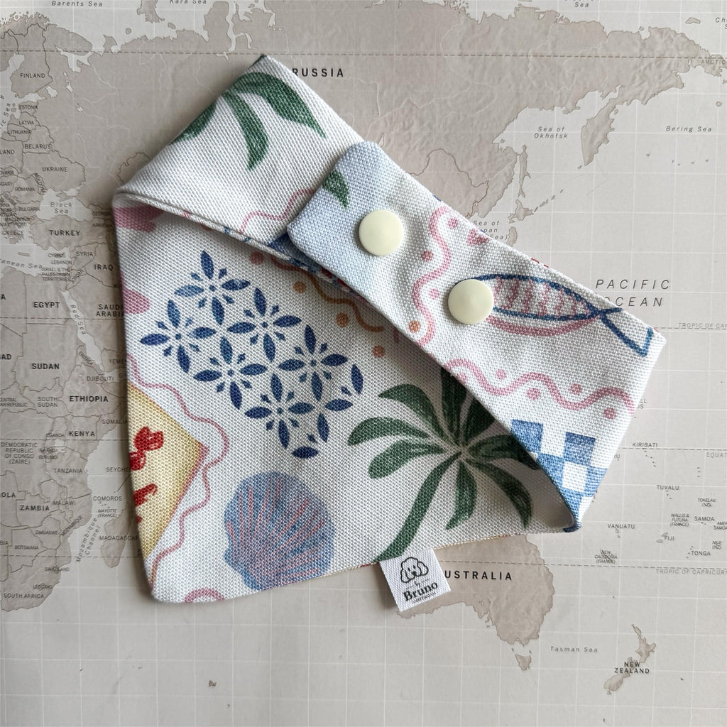 Back side of the white handmade dog bandana with pastel summer symbols like palm trees, crab and sea shell with plastic buttons on a world map background.