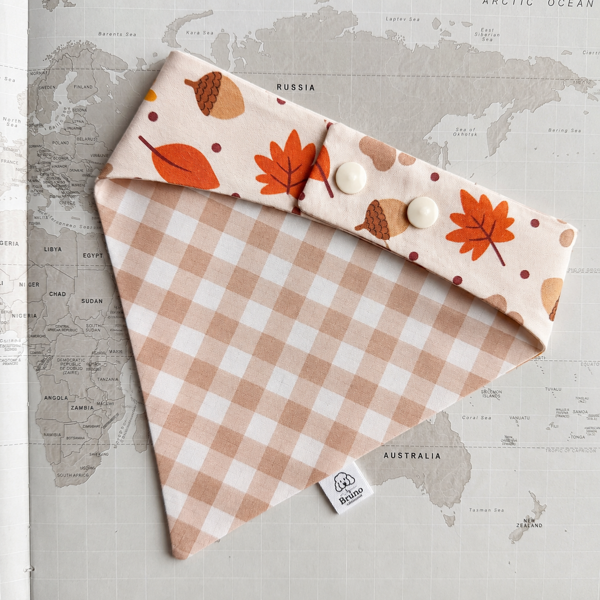 A dog bandana laid flat on a world map background. The bandana has a warm beige and white gingham pattern on the front and a matching top flap with an autumn leaf and acorn print, secured by two cream buttons.