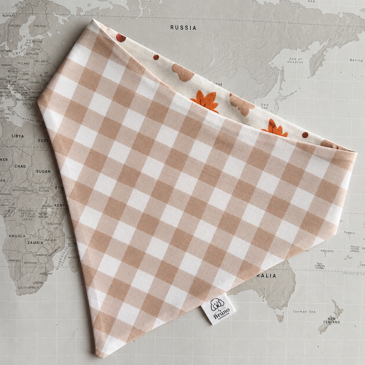 A dog bandana laid flat on a vintage-style world map. The bandana has a soft blush brown and white gingham pattern on the front, with a contrasting inner lining featuring orange leaves and cream dots.