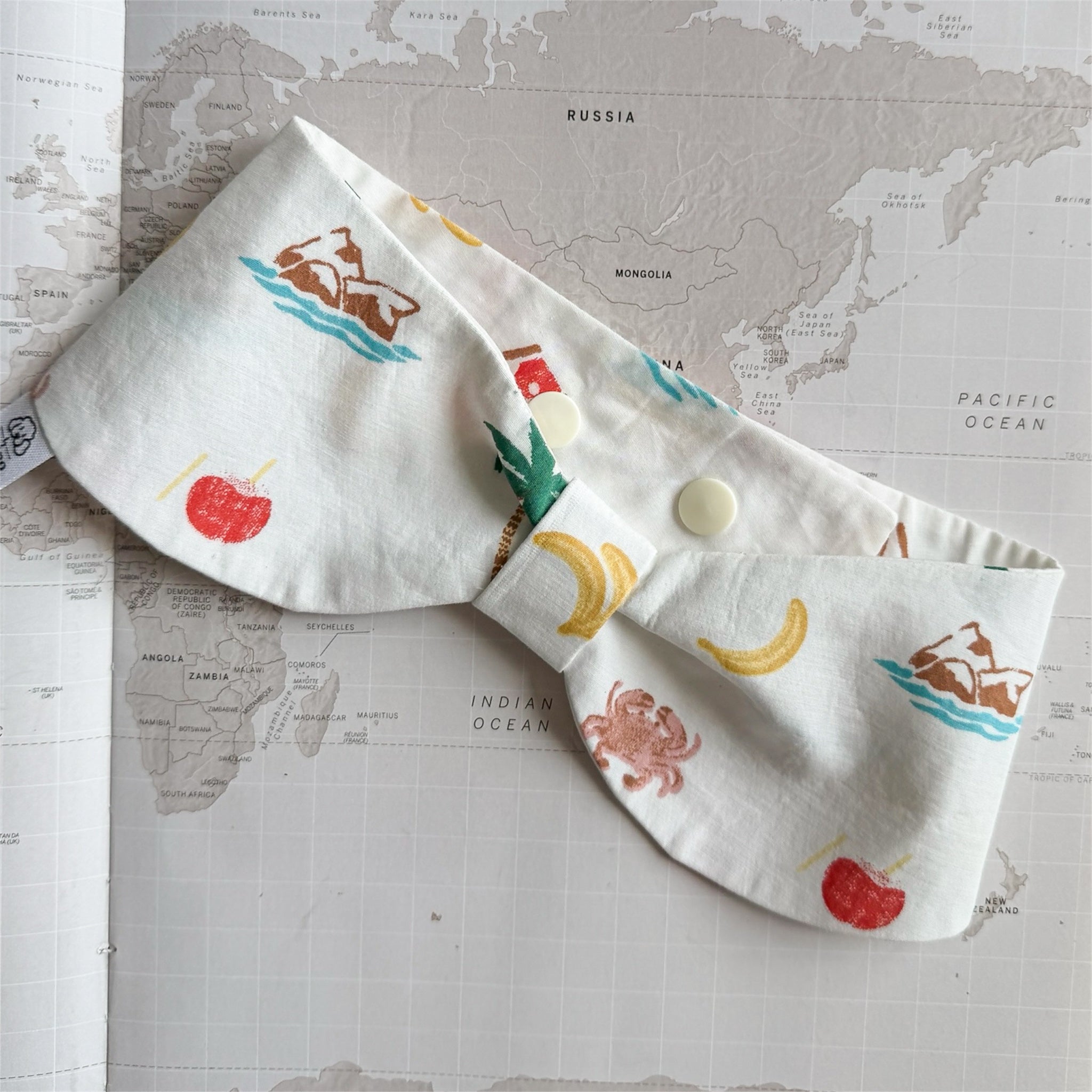 White handmade dog bowdana with summer symbols like crabs, palm trees, bananas on a world map background.