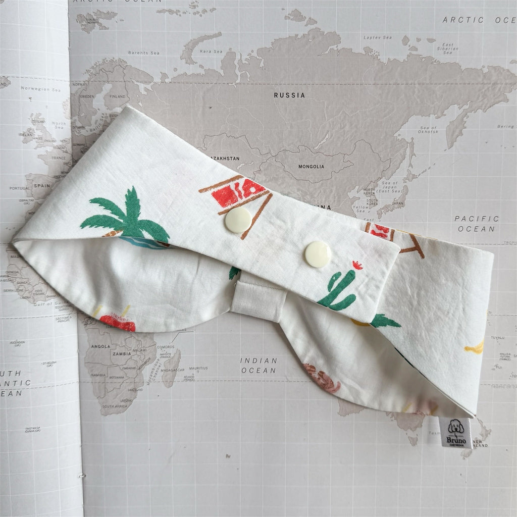 Back side of the white handmade dog bowdana with summer symbols like crabs, palm trees, bananas with plastic buttons on a world map background.