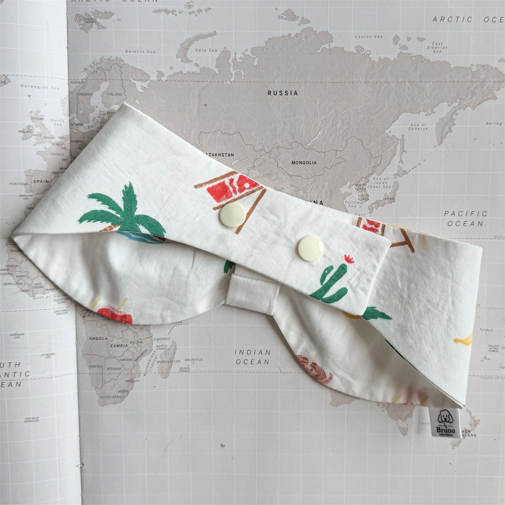 Back side of the white handmade dog bowdana with summer symbols like crabs, palm trees, bananas with plastic buttons on a world map background.